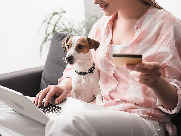 Frau mit einer rosa Bluse sitzt mit ihrem Hund auf dem Sofa und hat eine Kreditkarte in der Hand